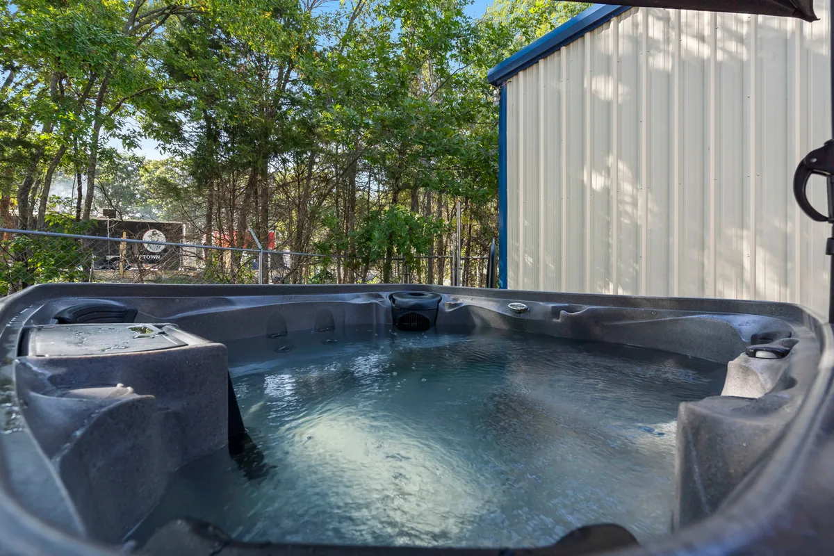 An inviting exterior shot of a private hot tub nestled among trees, bordered by a privacy fence and located just outside the main living areas. The tranquil setting hints at both group relaxation and quiet moments under the stars.