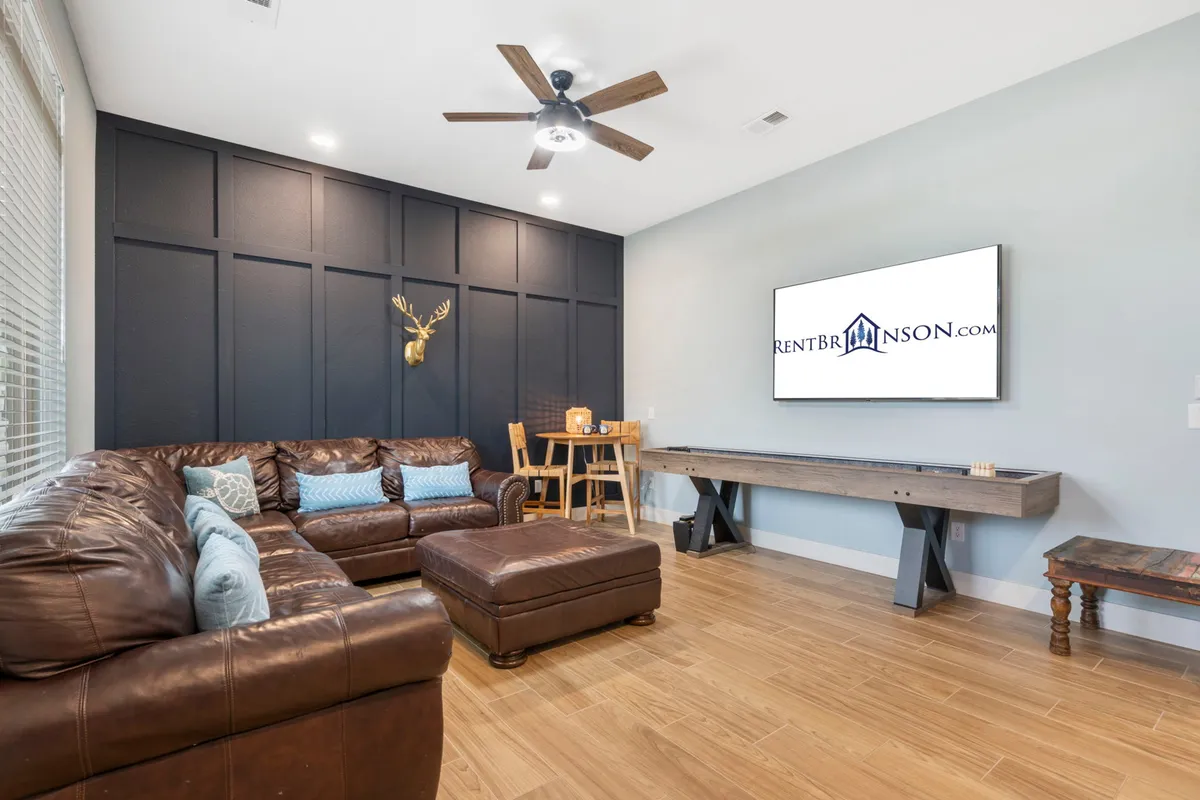 This image features a spacious living area with a leather sectional sofa, a large screen TV mounted on a wooden wall adorned with deer head decor, and a wooden table with chairs nearby. The ample seating and central entertainment area are ideal for group movie nights or board games.