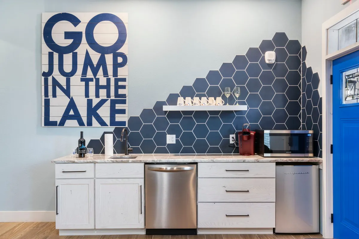 The interior features a bold blue wall art piece stating 'GO JUMP IN THE LAKE,' contemporary hexagonal tile backsplash, and a bright blue door. The playful decor reflects a lakeside lifestyle and encourages guests to embrace the water activities nearby, while the kitchen setting hints at social gatherings after exploring outdoors.