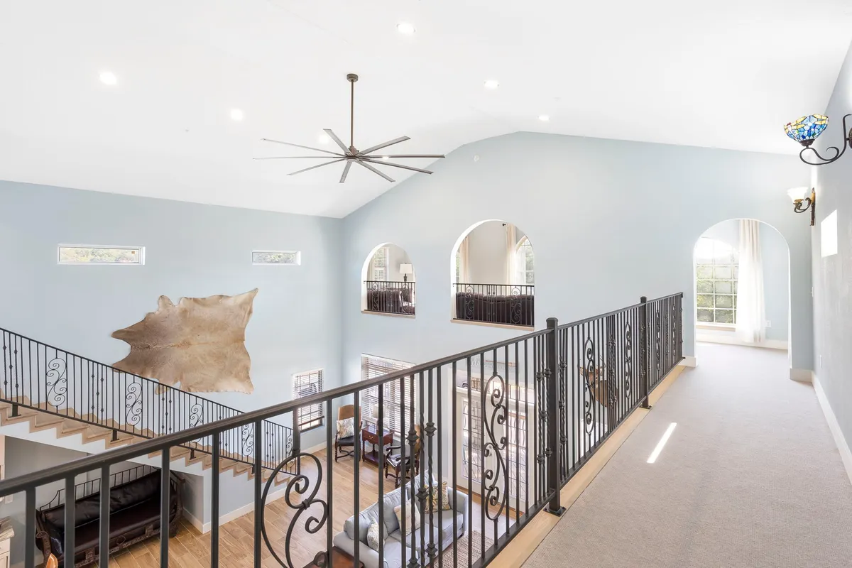 A bright, airy space with a large ceiling fan, decorative railing, abundant natural light from multiple windows, and elegant lighting fixtures. The open layout suggests a place for gathering and relaxing with loved ones.