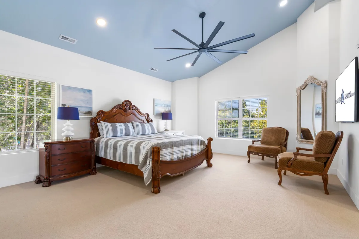 A serene king-sized bedroom featuring an ornate headboard, striped bedding, large ceiling fan, mirrored accent table, and comfortable armchairs. The room is flooded with natural light, creating a peaceful retreat ideal for relaxation or quiet moments.