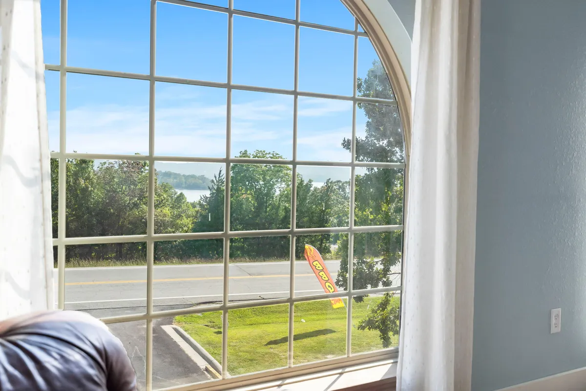 Through a large arched window, lush greenery and a clear blue sky are visible, blending the comfort of indoor spaces with an immediate connection to the outdoors. The inviting setup by the window suggests a space to relax and enjoy the view after a lakeside stroll or hike.