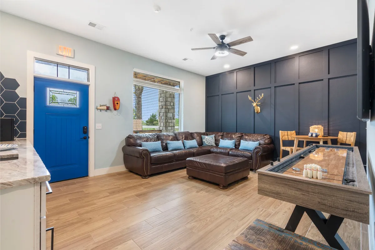 A welcoming interior featuring a blue front door, a leather sectional sofa, a wall-mounted deer head, and a dedicated game table. The space feels inviting and is clearly designed for casual gatherings, playing games, and enjoying time with friends or family.