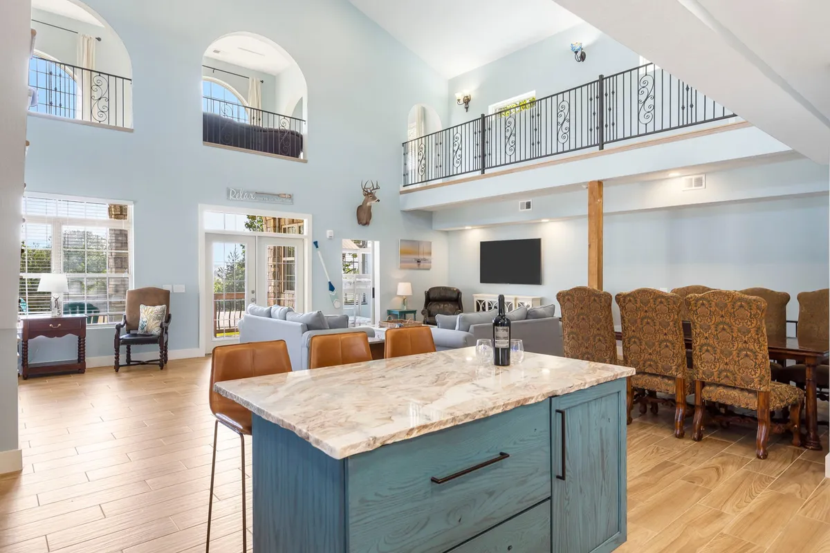 A bright, open-concept living space combining a kitchen island, dining area, living room with sofas, and a visible balcony above. Large windows let in natural light, and the area is set for family gatherings, entertaining guests, and relaxed evenings.