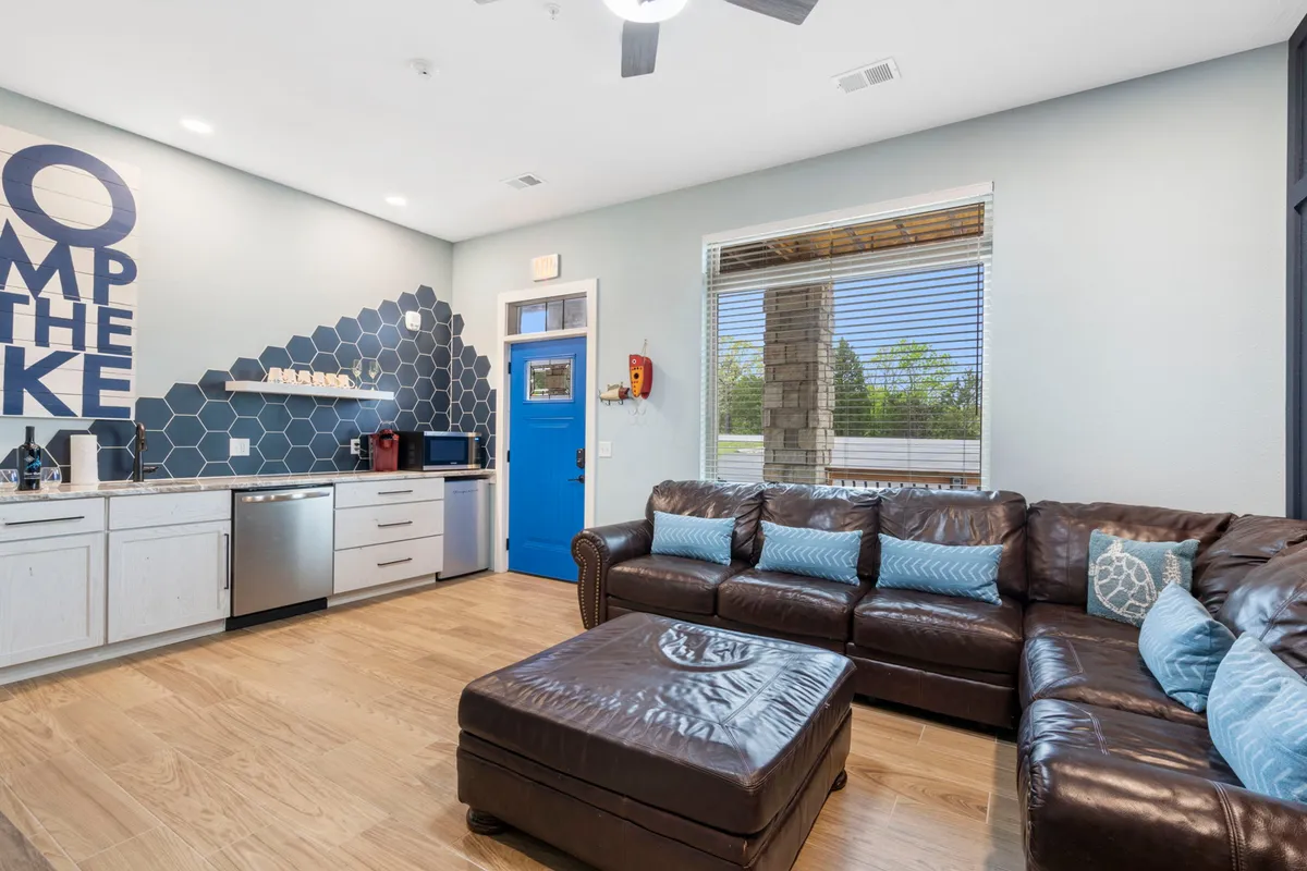 This image features a ground-floor kitchenette with stainless steel appliances, a blue accent wall displaying the phrase 'Jump the Lake,' a leather sectional sofa, and colorful decor accents. The overall scene is relaxed and welcoming, suggesting a space designed for prepping snacks, casual meals, and socializing before or after outdoor adventures.