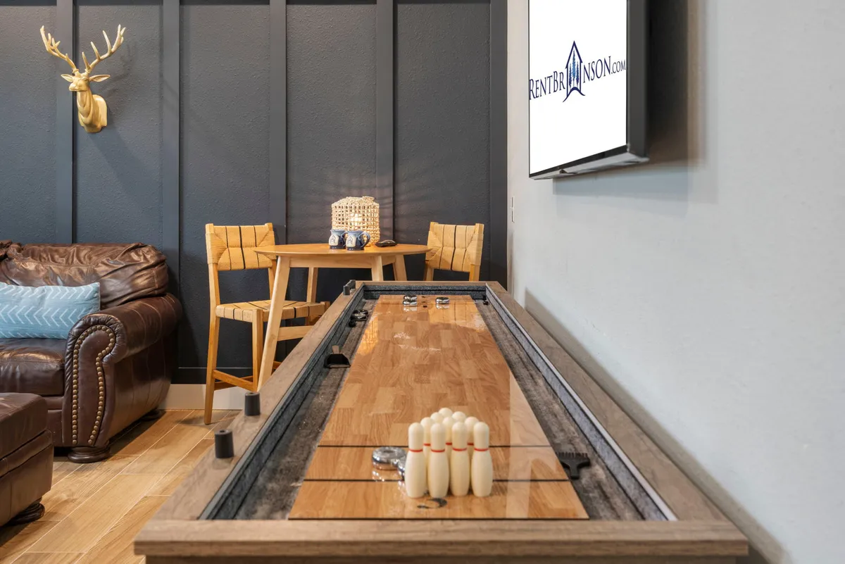 This image depicts a lively interior entertainment zone featuring a shuffleboard table, a leather couch, a small dining table, and a wall-mounted TV, accented with decorative antlers. The space is arranged for both relaxation and friendly competition, capturing the heart of the home's social hub.